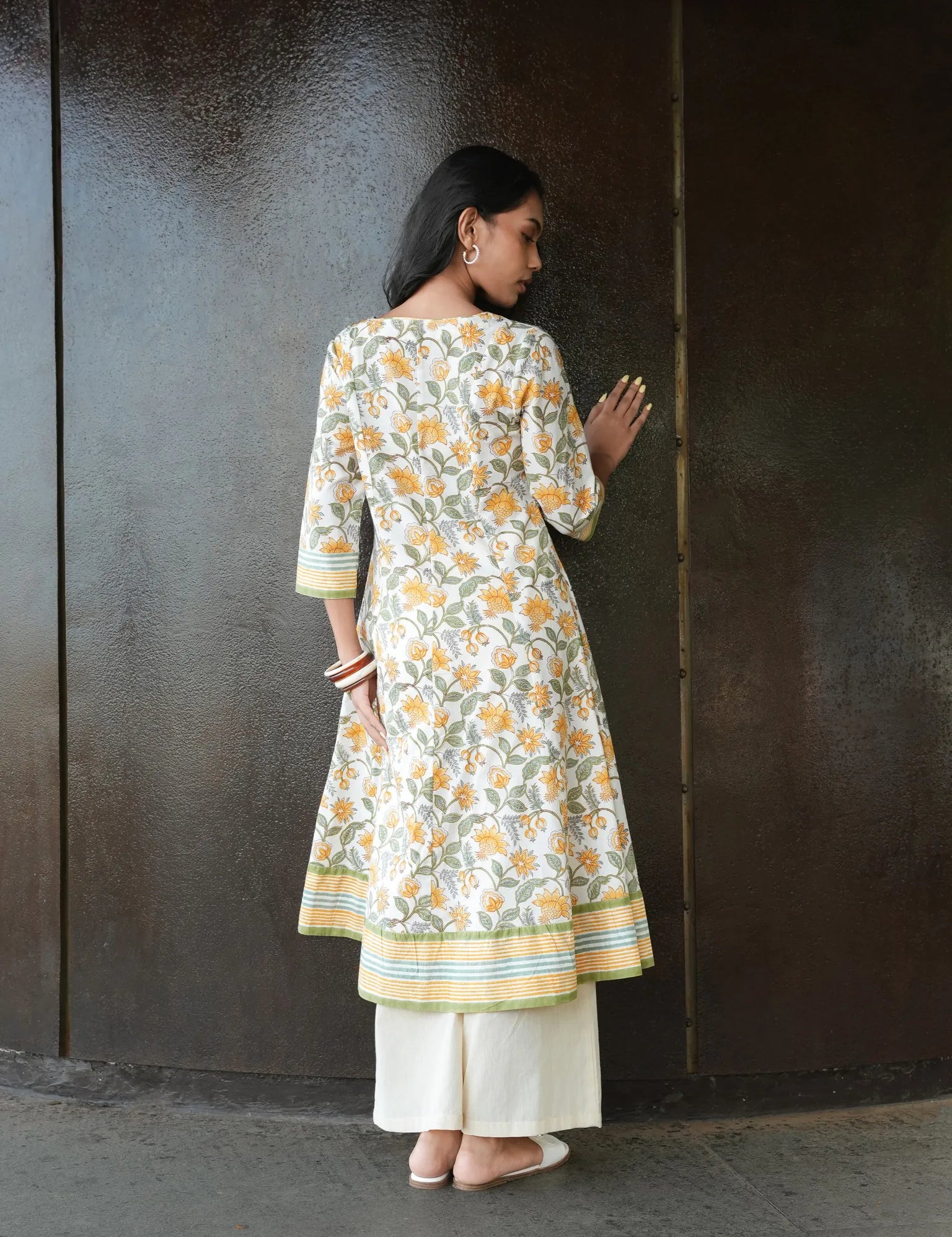 Woman wearing a floral kurta with white pants against a dark wall.