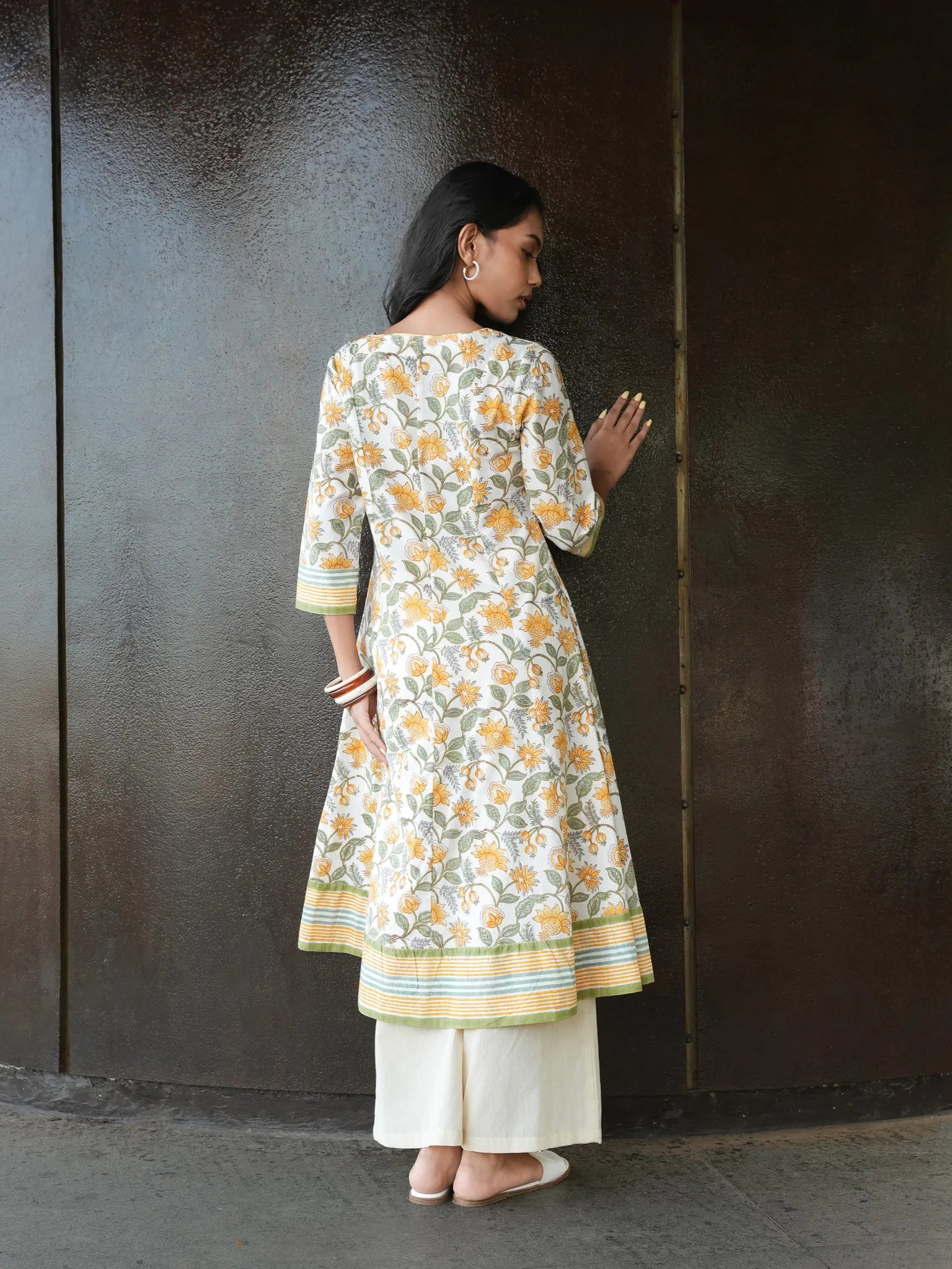 Woman wearing a floral kurta with white pants against a dark wall.