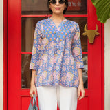 Woman wearing a blue floral blouse and white pants standing in front of a red door.