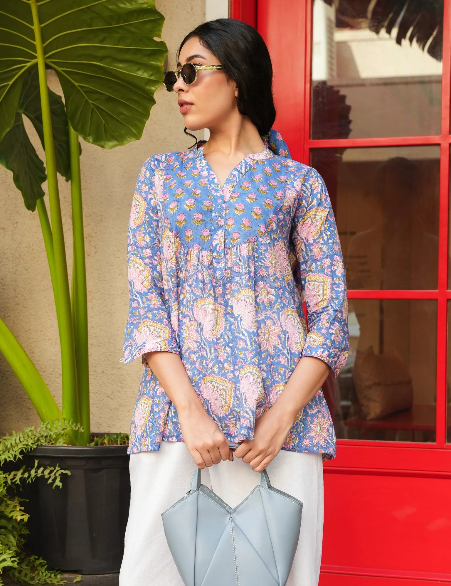 Woman wearing a blue patterned top holding a light blue handbag in front of a red door.