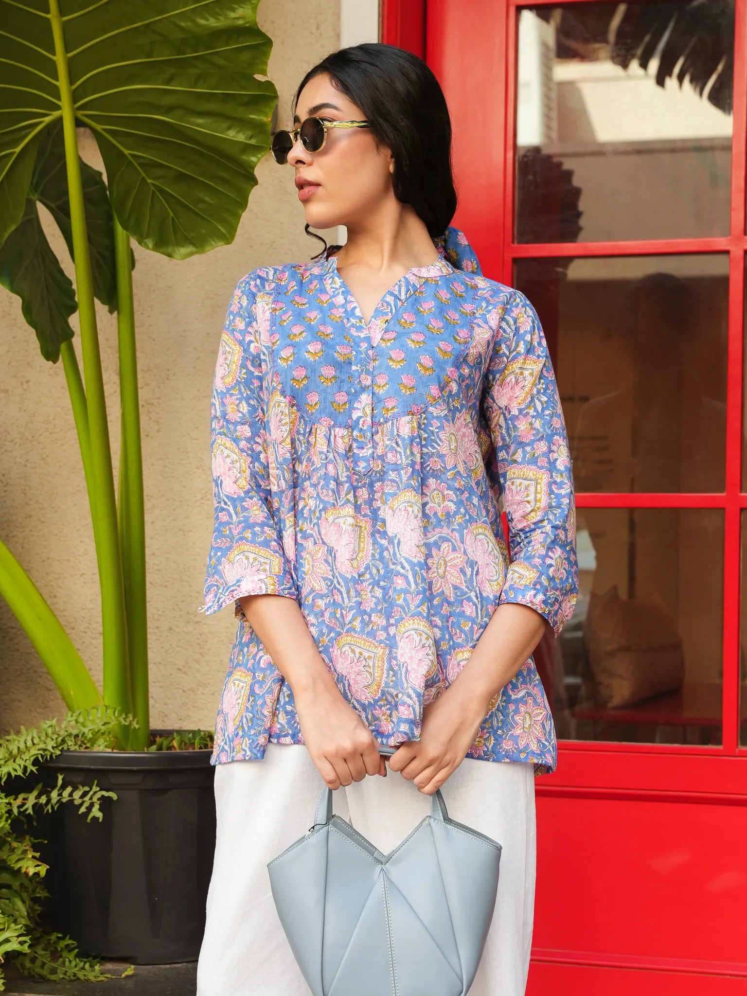 Woman wearing a blue patterned top holding a light blue handbag in front of a red door.