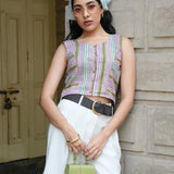 Woman wearing a striped sleeveless top and white skirt, holding a green handbag, standing against a wooden door.