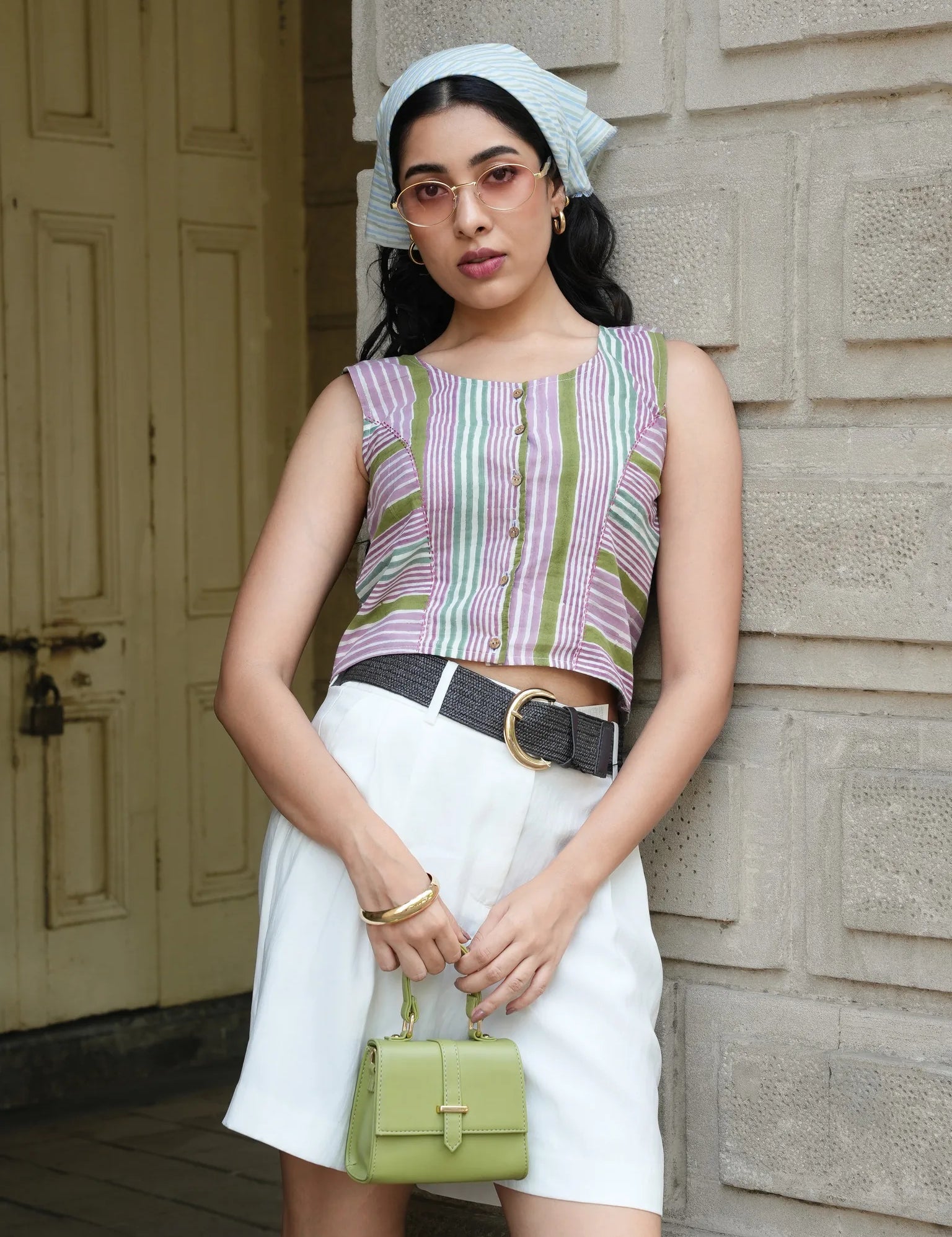 Woman wearing a striped sleeveless top and white skirt, holding a green handbag, standing against a wooden door.
