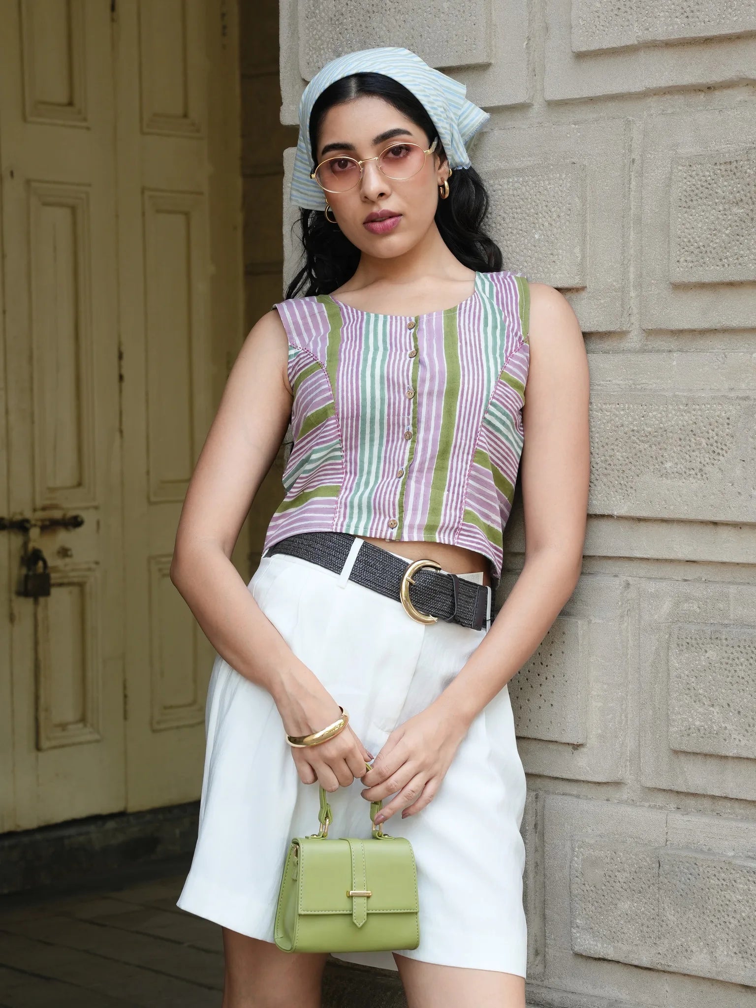 Woman wearing a striped sleeveless top and white skirt, holding a green handbag, standing against a wooden door.