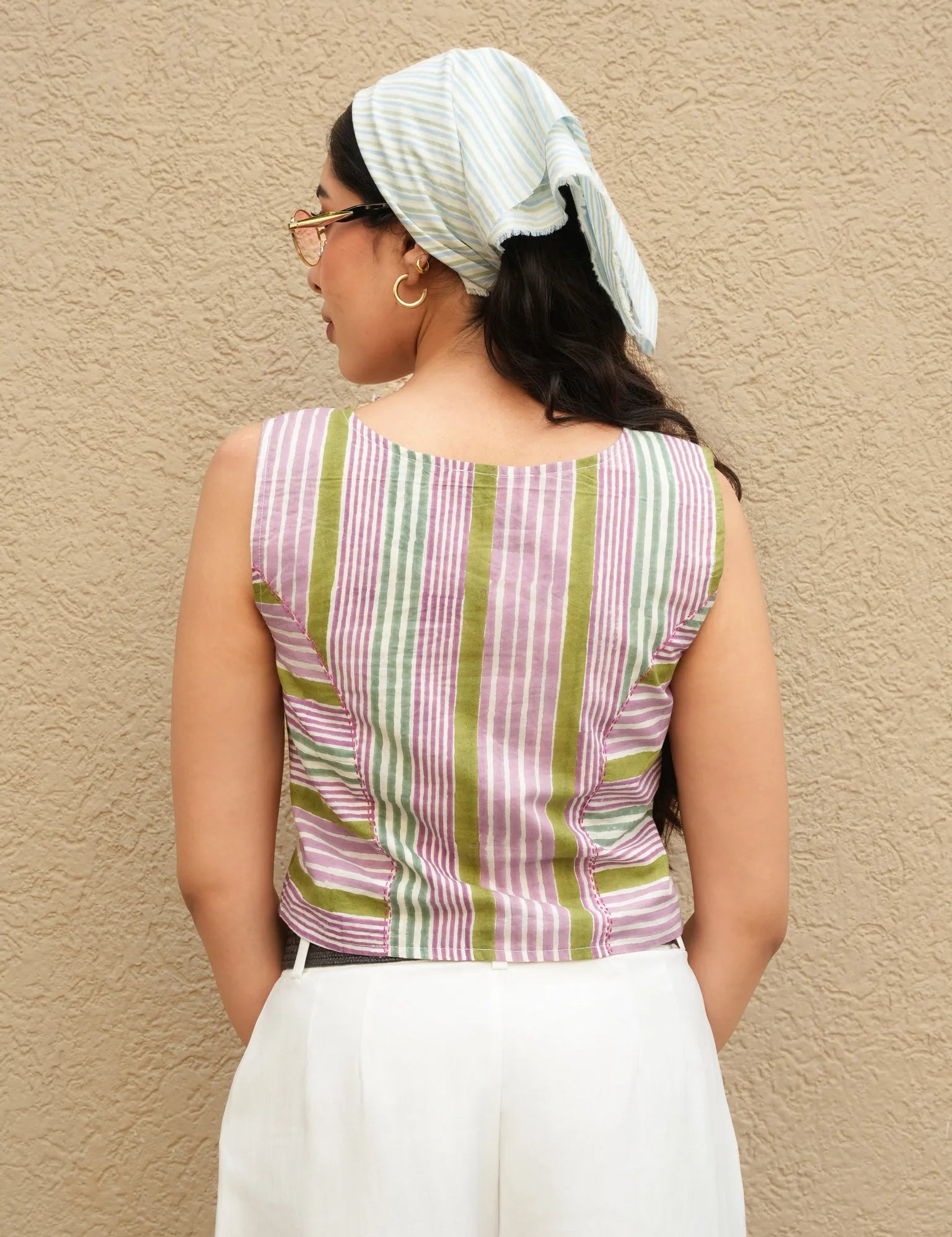 Woman wearing a striped sleeveless top and white pants against a beige wall.