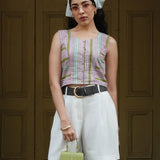 Woman wearing a striped sleeveless top and white shorts, holding a green handbag, against a wooden door background.