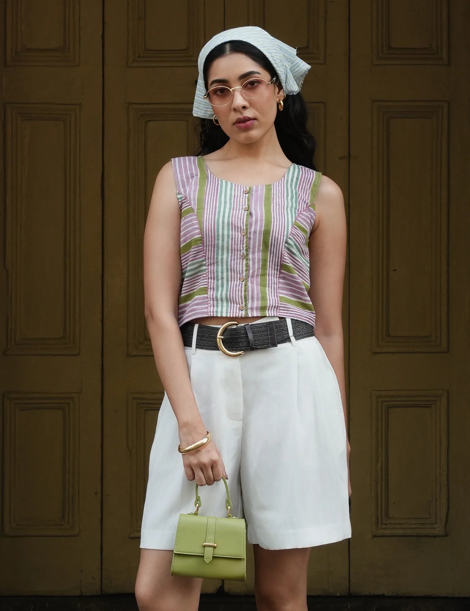 Woman wearing a striped sleeveless top and white shorts, holding a green handbag, against a wooden door background.