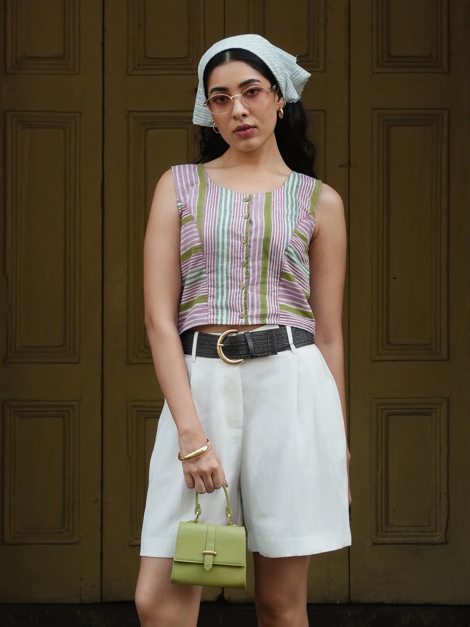 Woman wearing a striped sleeveless top and white shorts, holding a green handbag, against a wooden door background.