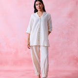 A woman modeling a two-piece white kurta and trouser set against a textured pink studio backdrop.