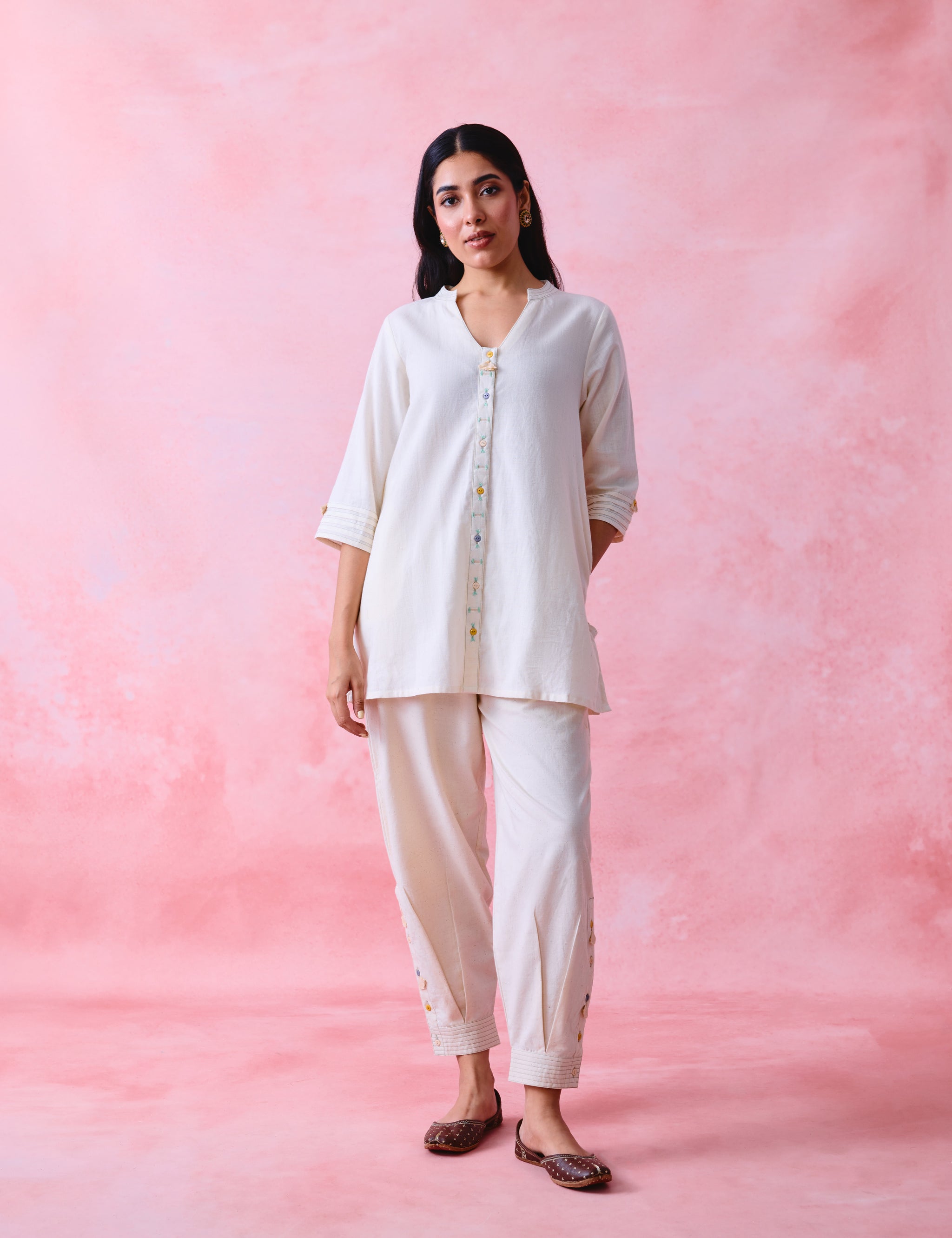 A woman modeling a two-piece white kurta and trouser set against a textured pink studio backdrop.