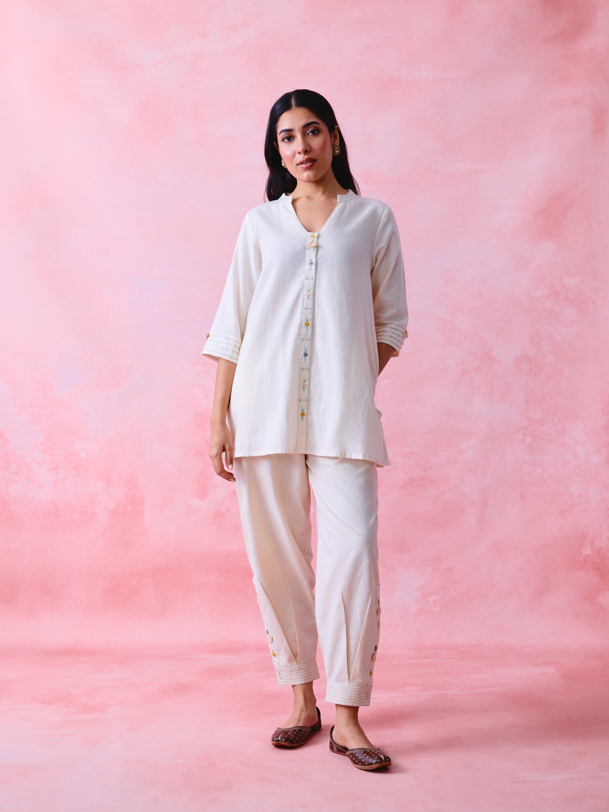 A woman modeling a two-piece white kurta and trouser set against a textured pink studio backdrop.