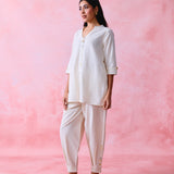 A woman modeling a cream-colored two-piece kurta and pant set against a textured pink studio backdrop.