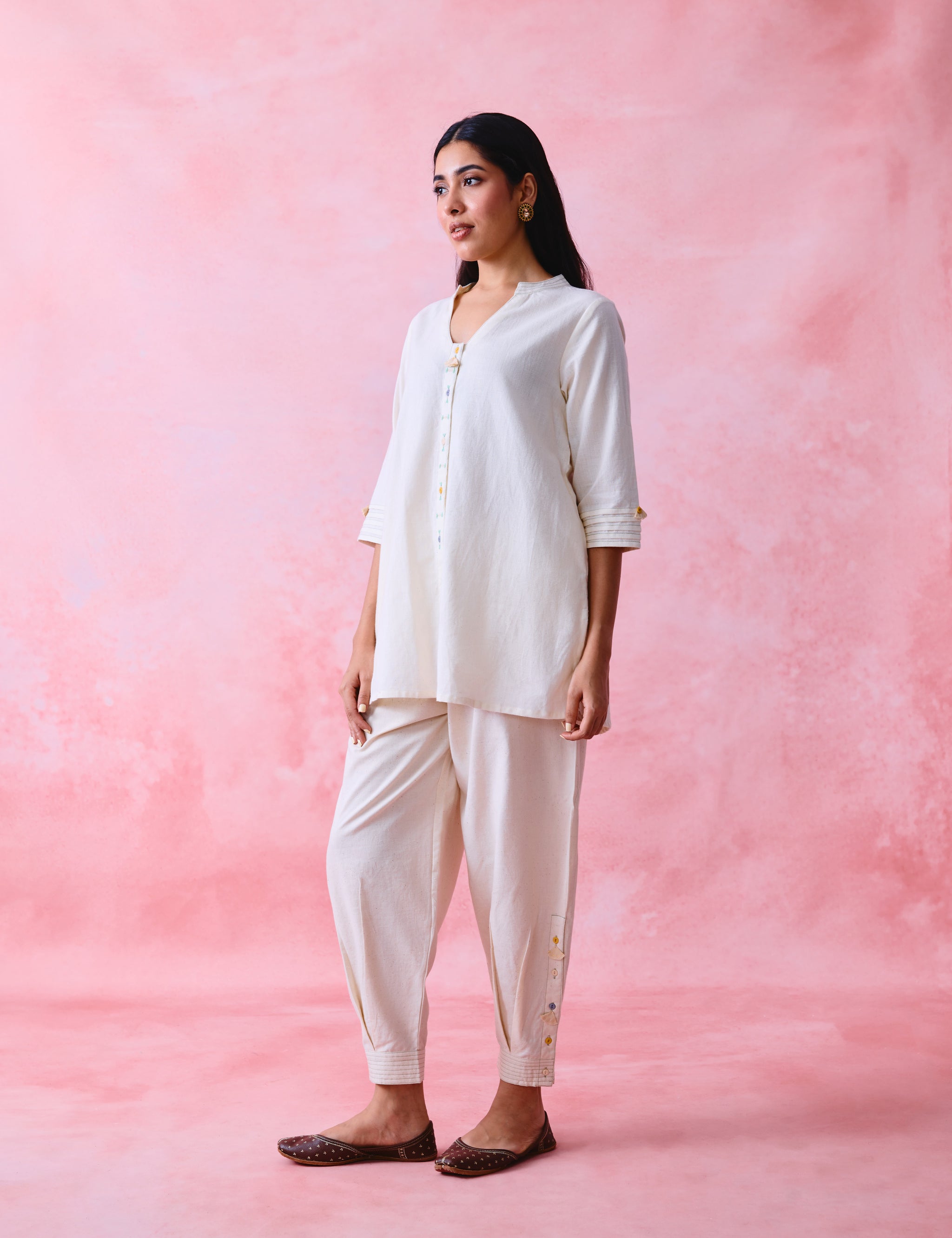 A woman modeling a cream-colored two-piece kurta and pant set against a textured pink studio backdrop.
