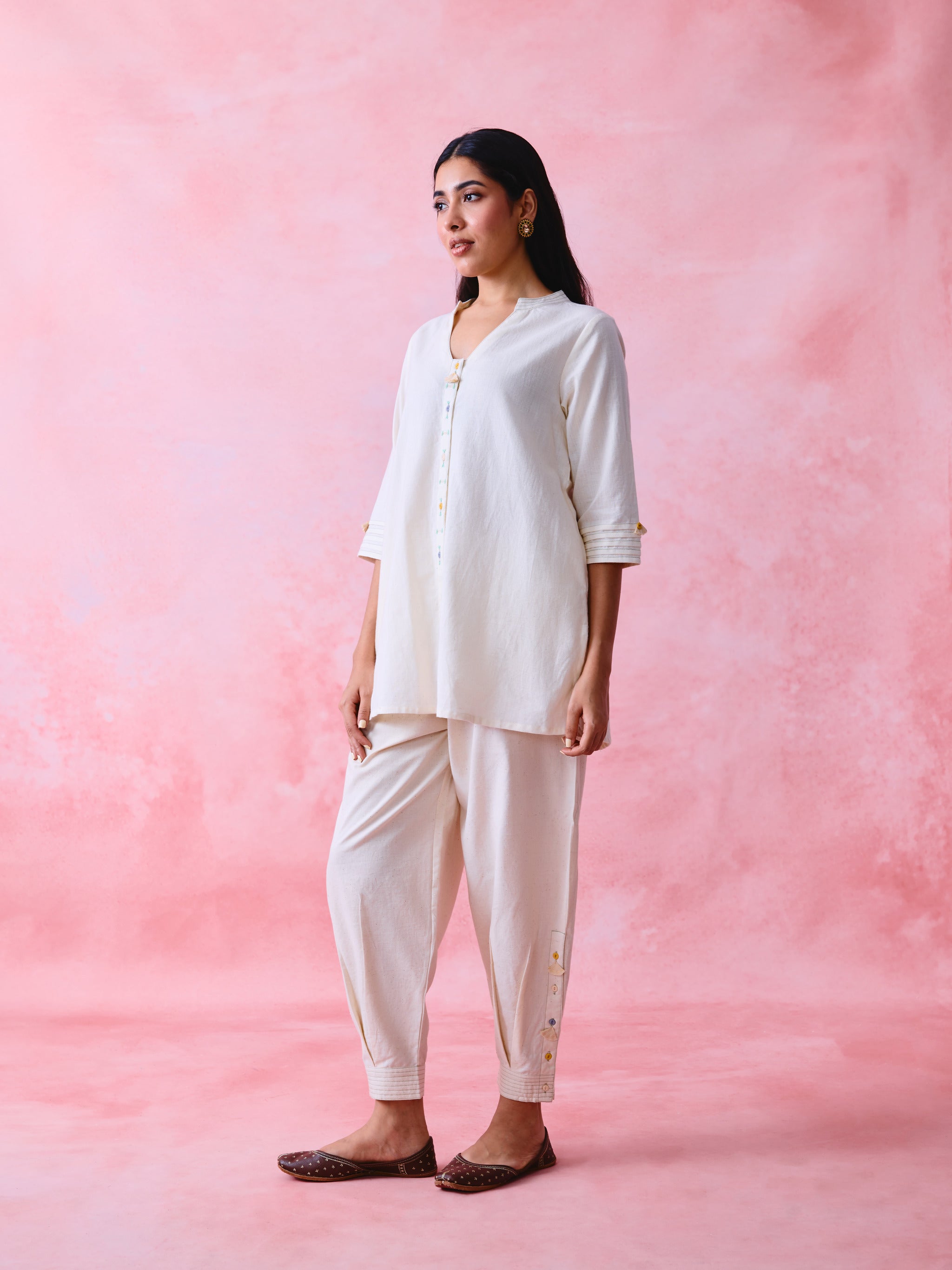 A woman modeling a cream-colored two-piece kurta and pant set against a textured pink studio backdrop.